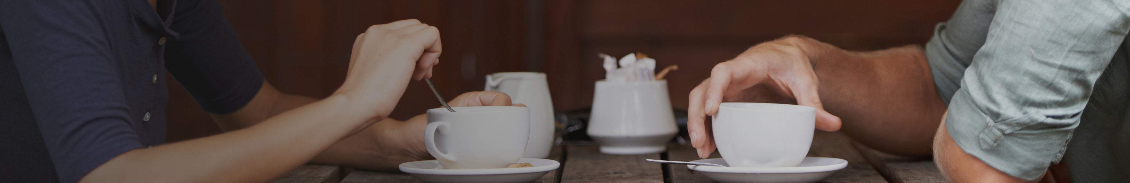 Two people sitting across from each other at a wooden table, each holding a white cup of coffee or tea.