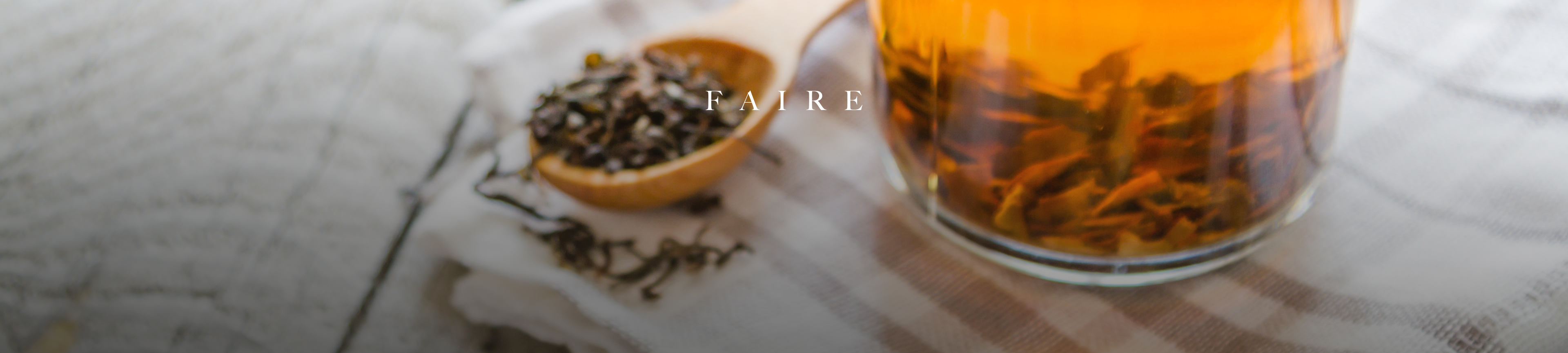A close-up shot shows a wooden spoon filled with loose-leaf tea next to a glass of brewed amber-colored tea. The word "F A I R E" is subtly overlaid on the image in white text.