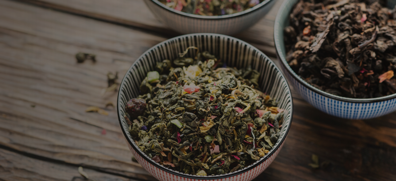 Assorted dried loose leaf teas in colorful ceramic bowls placed on a rustic wooden surface.
